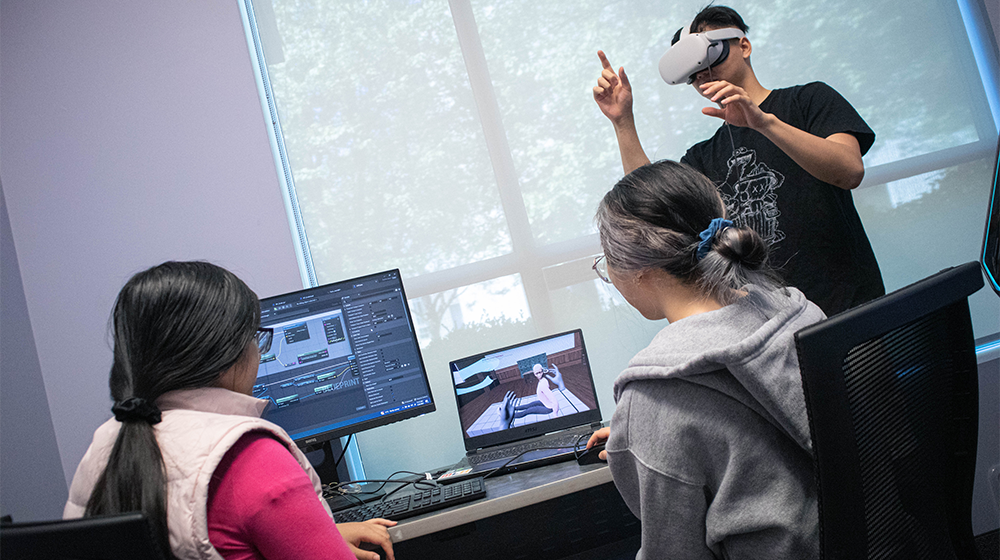 Three people working on a computer, one of them is wearing a VR helmet